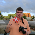 Jakob steht mit einer Kamera und einer Dänemarkflagge vor einem Bahnübergang.